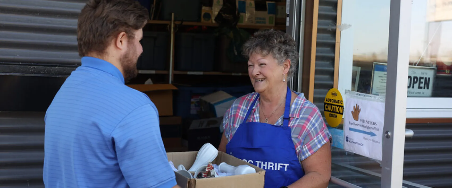 A man donating a box of pre-owned goods to an MCC Thrift shop volunteer.